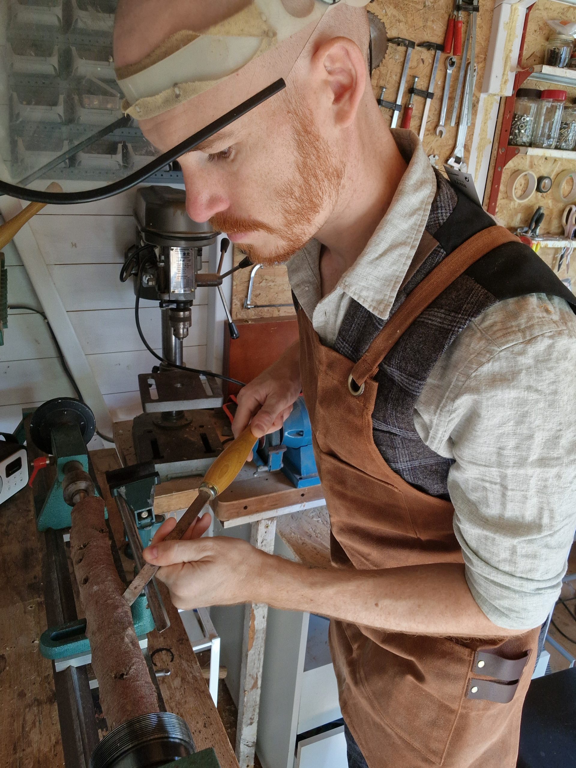 Craftsman shaping a wooden wand on a lathe in a workshop, wearing apron and face shield